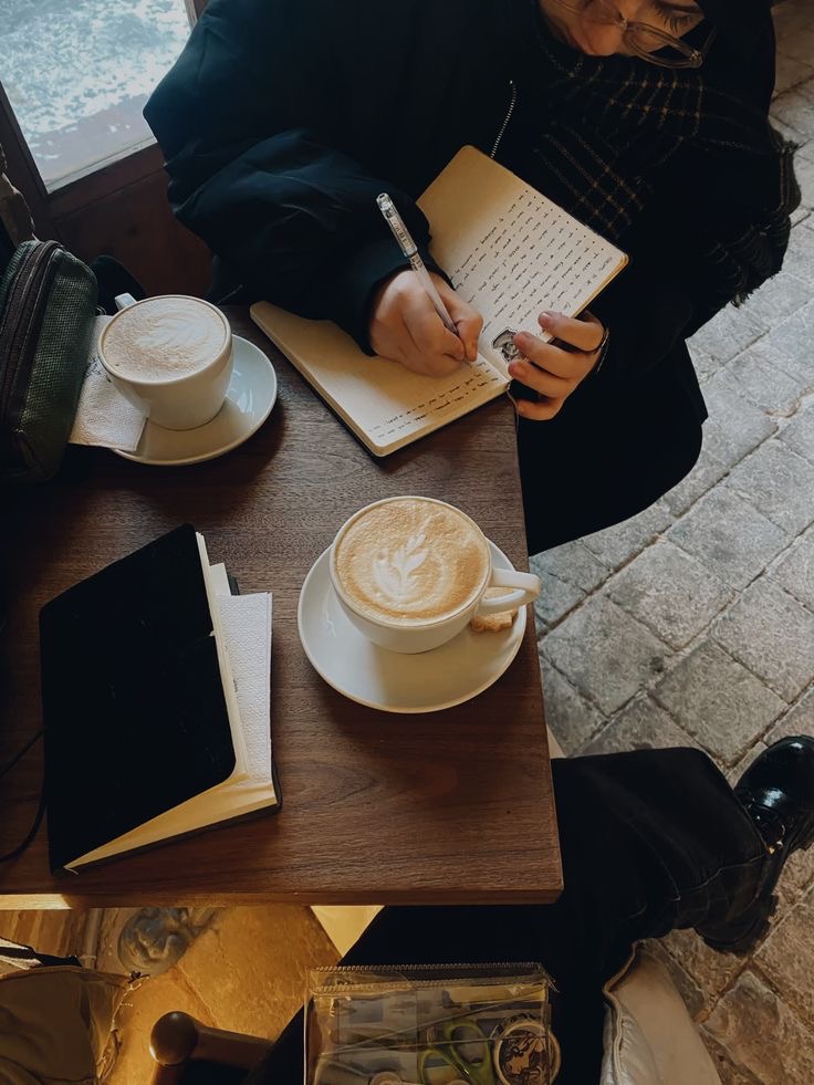 Two coffees and an open notebook on a café table, a woman writing during a first coaching session.